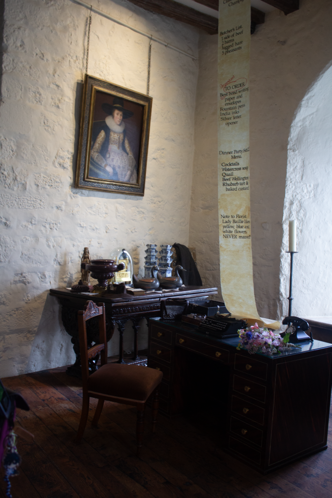 Intérieur d'une salle du château de Leeds. Portrait d'une femme sur un mur blanc. Un bureau avec machine à écrire et téléphone anciens. Long parchemin pendu au plafond descend sur le bureau